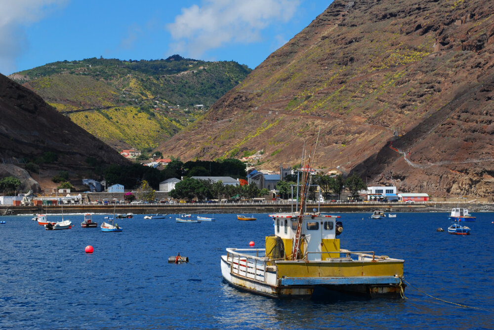 a yellow and white fishing boat tied to a mooring in the bay with a number of smaller boats around and a brown island beyond with very steep-sided mountains and a town nestled in the valley