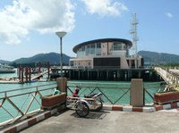 Thailand-Phuket-Ao_Chalong_Pier railings on a pier with a trolley parked next to them - blue water behind and the other side of the water a round building with glass and a flat roof