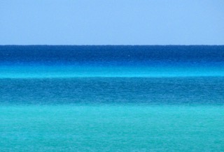horizontal lines of bright blue sea of varying shades topped by a pale blue solid colour sky not a cloud in sight