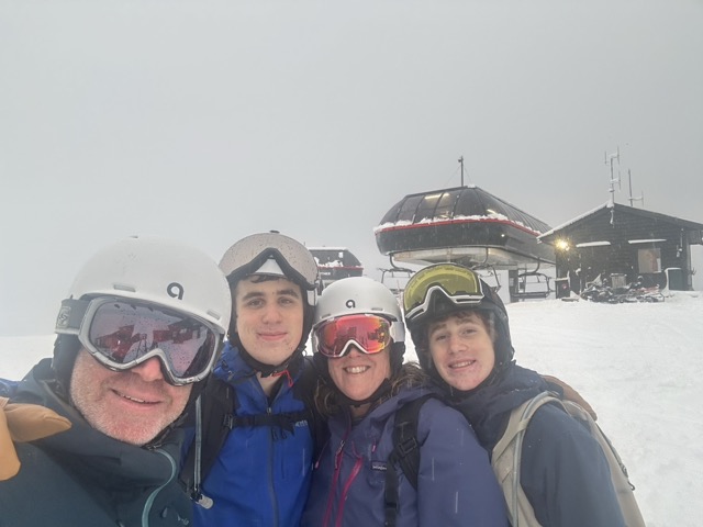 4 smiling people in cold weather gear and helmets with a chair lift behind them
